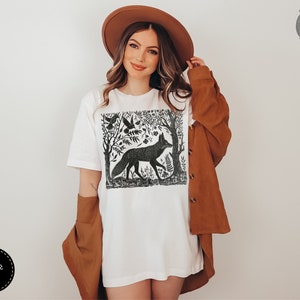 May include: White t-shirt with a black and white illustration of a fox in a forest setting. The illustration includes trees, birds, and flowers. The text "Forest Fables" is in the top right corner of the image.