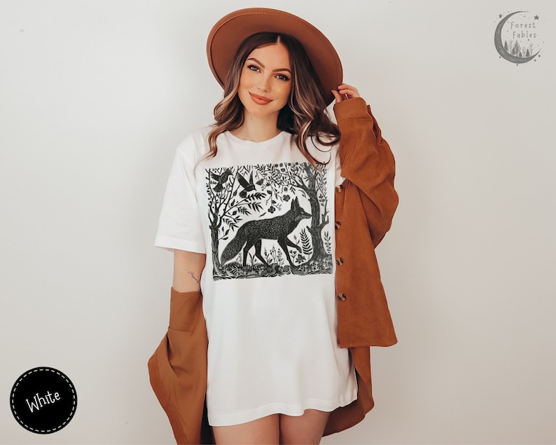 May include: White t-shirt with a black and white illustration of a fox in a forest setting. The illustration includes trees, birds, and flowers. The text "Forest Fables" is in the top right corner of the image.