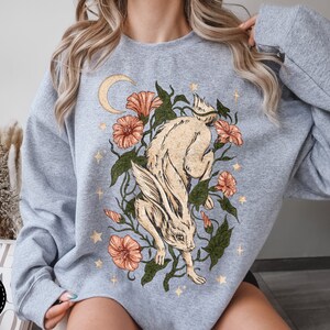 May include: A light gray sweatshirt with a graphic print of a white rabbit running through a field of flowers and stars. The flowers are orange, pink, and red. The rabbit has a crescent moon behind it.