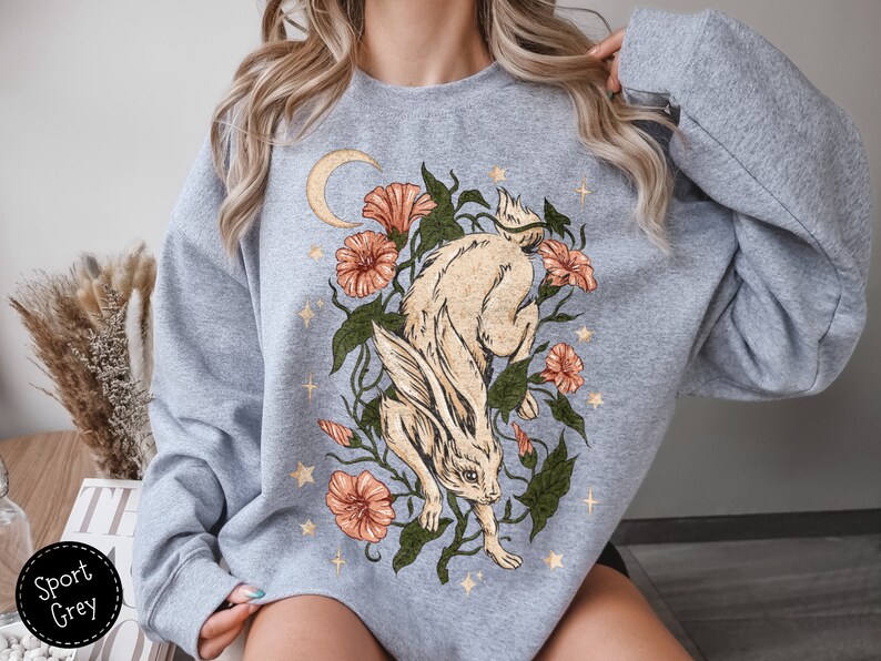 May include: A light gray sweatshirt with a graphic print of a white rabbit running through a field of flowers and stars. The flowers are orange, pink, and red. The rabbit has a crescent moon behind it.