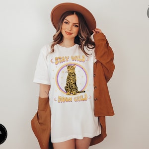 May include: White t-shirt with a graphic of a cat sitting with the text "Stay Wild Moon Child" in a retro font.
