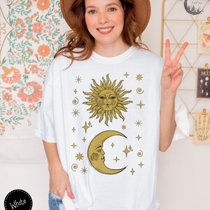 Puede incluir: Camiseta blanca con un dise&ntilde;o de sol y luna dorados. El sol y la luna tienen caras y est&aacute;n rodeados de estrellas.