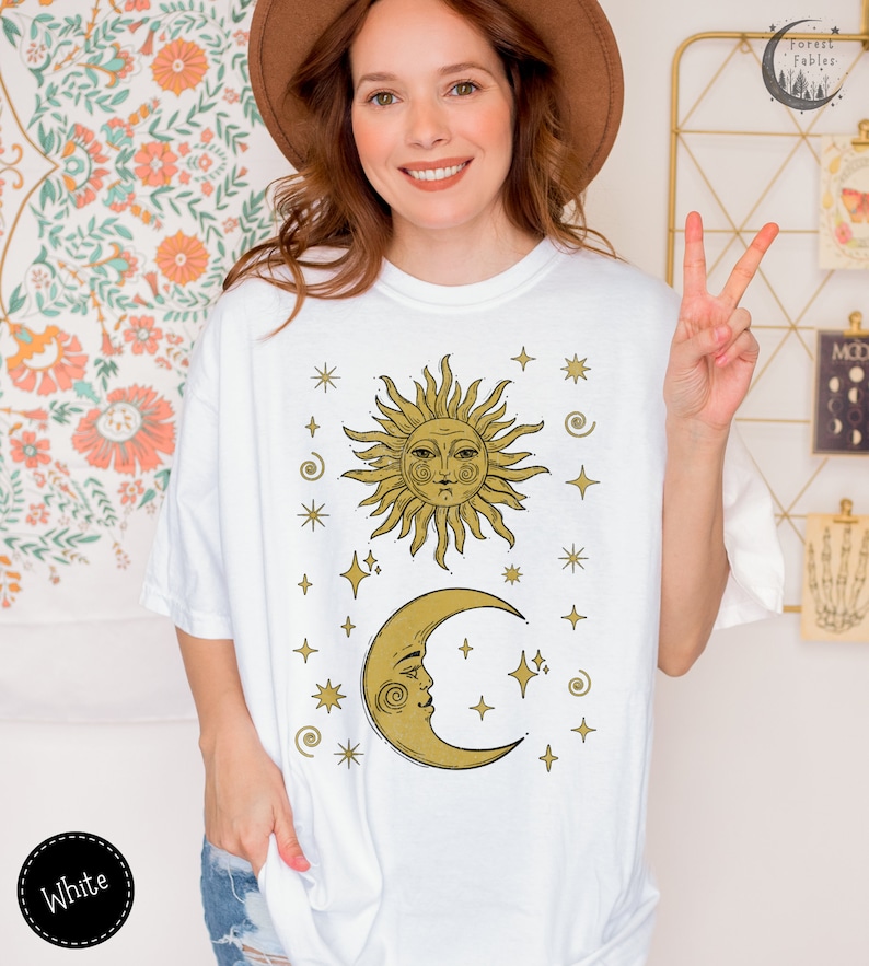 Puede incluir: Camiseta blanca con un dise&ntilde;o de sol y luna dorados. El sol y la luna tienen caras y est&aacute;n rodeados de estrellas.
