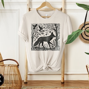 May include: A beige t-shirt with a black and white illustration of a fox in a forest scene. The illustration is a woodblock print style.