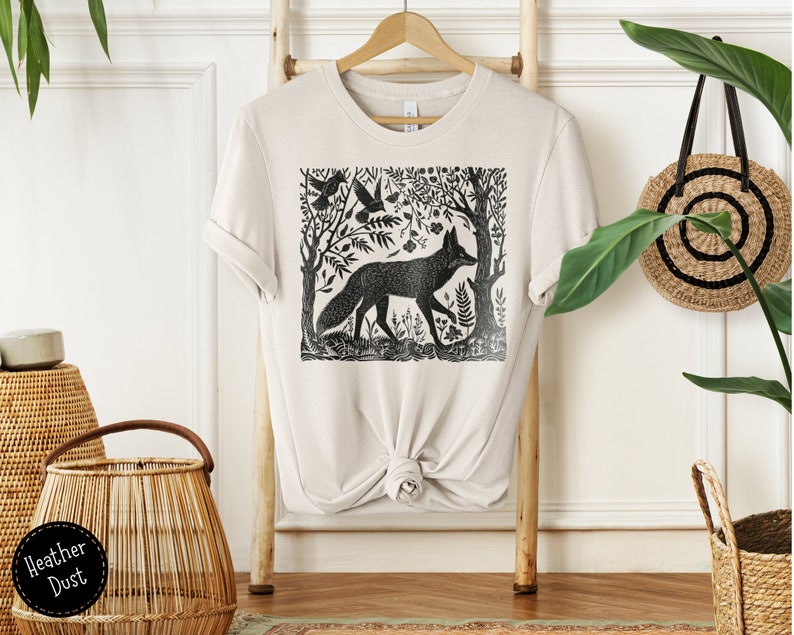 May include: A beige t-shirt with a black and white illustration of a fox in a forest scene. The illustration is a woodblock print style.