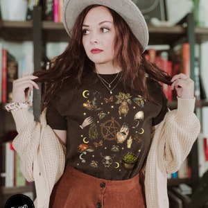 Puede incluir: Una camiseta negra con un estampado gr&aacute;fico de varios s&iacute;mbolos de bruja, incluyendo una luna creciente, un pentagrama, una mano con un ojo, un caldero y una calavera. La camiseta la lleva puesta una persona con el pelo casta&ntilde;o y un sombrero gris.