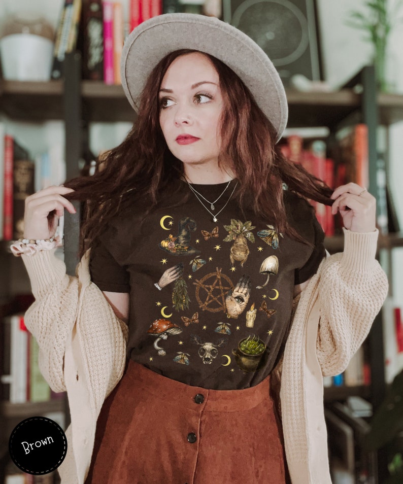 Puede incluir: Una camiseta negra con un estampado gr&aacute;fico de varios s&iacute;mbolos de bruja, incluyendo una luna creciente, un pentagrama, una mano con un ojo, un caldero y una calavera. La camiseta la lleva puesta una persona con el pelo casta&ntilde;o y un sombrero gris.