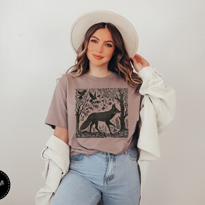 May include: A woman wearing a mauve t-shirt with a black and white graphic print of a fox in a forest scene. The t-shirt has a crew neck and short sleeves. The woman is also wearing a white hat and a white cardigan.