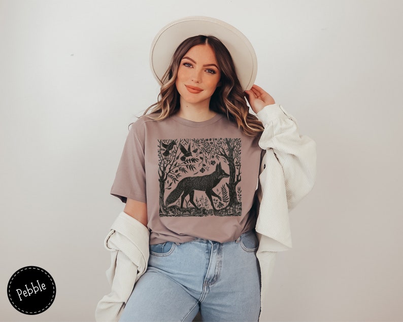 May include: A woman wearing a mauve t-shirt with a black and white graphic print of a fox in a forest scene. The t-shirt has a crew neck and short sleeves. The woman is also wearing a white hat and a white cardigan.
