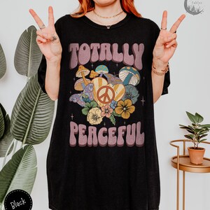 May include: Black oversized t-shirt with a retro graphic design featuring the text "Totally Peaceful" in a groovy font. The graphic also includes a peace sign, mushrooms, and flowers.