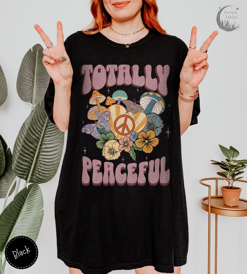 May include: Black oversized t-shirt with a retro graphic design featuring the text "Totally Peaceful" in a groovy font. The graphic also includes a peace sign, mushrooms, and flowers.