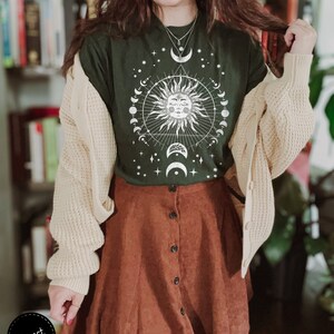 K&ouml;nnte beinhalten: Ein gr&uuml;nes T-Shirt mit einem wei&szlig;en Sonnen- und Monddesign. Das Design zeigt eine Sonne mit einem Gesicht, umgeben von einem Kreis aus Monden und Sternen. Das Shirt wird mit einem braunen Cordrock kombiniert.