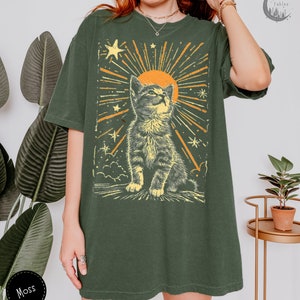 May include: A green oversized t-shirt with a vintage-style graphic of a cat looking up at a sun with stars and rays of light. The text "Forest Fables" is in the top right corner of the image.