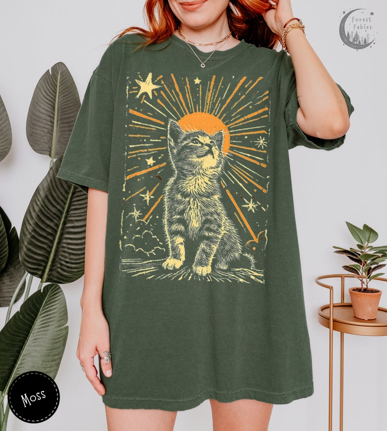 May include: A green oversized t-shirt with a vintage-style graphic of a cat looking up at a sun with stars and rays of light. The text "Forest Fables" is in the top right corner of the image.