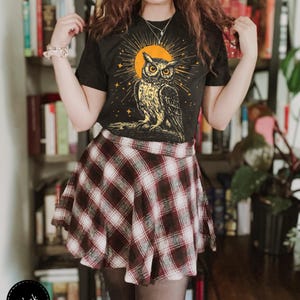 May include: Black t-shirt featuring a graphic of an owl in front of a yellow sun and stars. The owl is surrounded by a circle of light. The shirt is being worn by a person with long brown hair and a plaid skirt.