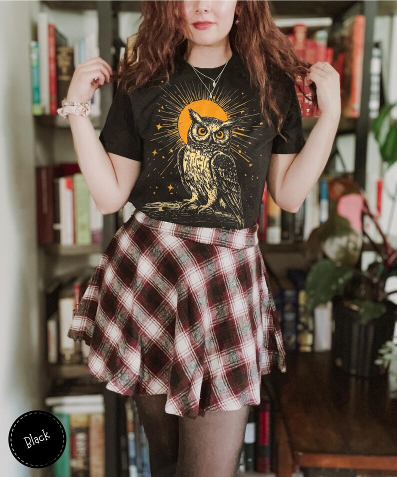 May include: Black t-shirt featuring a graphic of an owl in front of a yellow sun and stars. The owl is surrounded by a circle of light. The shirt is being worn by a person with long brown hair and a plaid skirt.