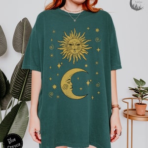 Puede incluir: Una camiseta verde azulada con un dise&ntilde;o de sol y luna dorado. El sol y la luna tienen caras y est&aacute;n rodeados de estrellas. El texto "Forest Fables" est&aacute; en la esquina superior derecha de la imagen.