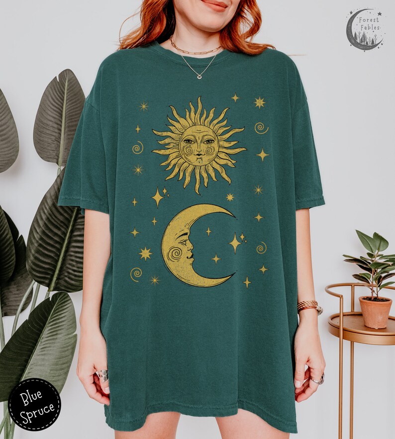 Puede incluir: Una camiseta verde azulada con un dise&ntilde;o de sol y luna dorado. El sol y la luna tienen caras y est&aacute;n rodeados de estrellas. El texto "Forest Fables" est&aacute; en la esquina superior derecha de la imagen.