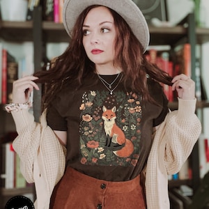 May include: A brown t-shirt featuring a fox illustration surrounded by flowers. The fox is depicted with orange and white fur, sitting amidst a floral design. The shirt is paired with a brown skirt and a beige cardigan.