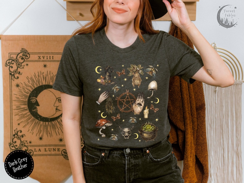 Puede incluir: Camiseta de color gris oscuro jaspeado con un estampado gr&aacute;fico de una escena de brujer&iacute;a con setas, un pentagrama, una mano con un ojo y otros elementos m&iacute;sticos. El texto "Forest Fables" es visible en la esquina superior derecha de la imagen.