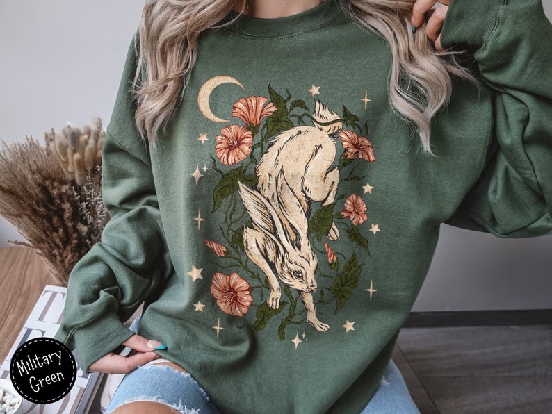 May include: A green sweatshirt with a white rabbit illustration surrounded by flowers and stars. The rabbit is jumping and has a crescent moon behind it.