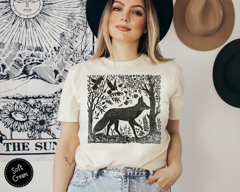 May include: A white t-shirt with a black and white illustration of a fox in a forest setting. The illustration includes trees, birds, and flowers.