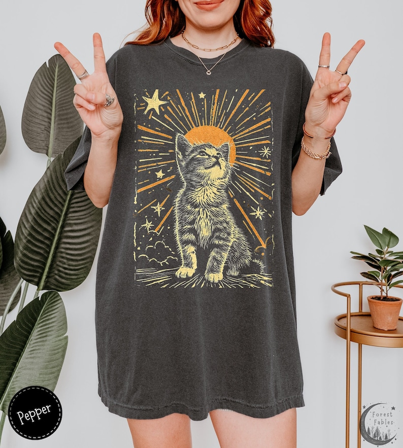 May include: A black oversized t-shirt with a graphic print of a yellow cat looking up at a yellow sun with yellow stars and rays of light. The cat is surrounded by clouds. The text "Forest Fables" is in the bottom right corner of the image.