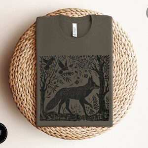 May include: A dark green t-shirt with a black and white illustration of a fox in a forest setting. The illustration includes trees, leaves, and birds. The text "Forest Fables" is in the upper right corner of the image.