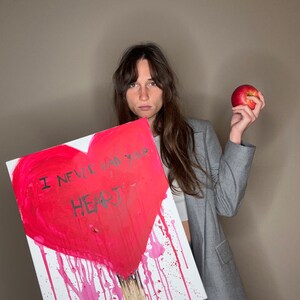 Può includere: Una donna tiene in mano un dipinto su tela con un cuore rosso dipinto. Il cuore ha le parole "I never had your heart" scritte su di esso. La donna tiene una mela rossa nella sua altra mano.