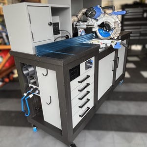 May include: A custom-built woodworking station with a black and gray frame, featuring a miter saw, cabinets, drawers, and a dust collection system. The station includes a blue and silver miter saw, and is mounted on wheels for mobility.