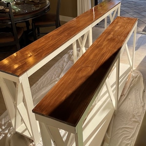 Puede incluir: Dos mesas consola de madera pintadas de blanco con una tapa de madera marrón. Las mesas tienen un diseño en forma de X en la parte delantera y los lados.