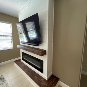 May include: A white shiplap wall with a mounted flat screen television above a dark wood mantel and a black electric fireplace. The fireplace is on a raised platform with a dark wood base.