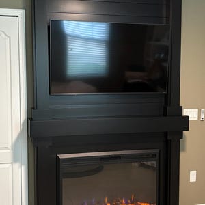 Puede incluir: Un marco de chimenea negro de suelo a techo con un diseño de paneles horizontales. La estructura incluye una chimenea eléctrica integrada con brasas de color naranja brillante en la base y un televisor de pantalla plana montado encima.