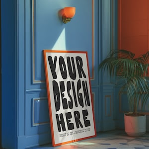 May include: A white poster with a black and orange frame, leaning against a blue wall. The poster says "YOUR DESIGN HERE" in large, black, stylized letters. The poster is in a room with a light blue wall, a light orange wall, and a potted plant.