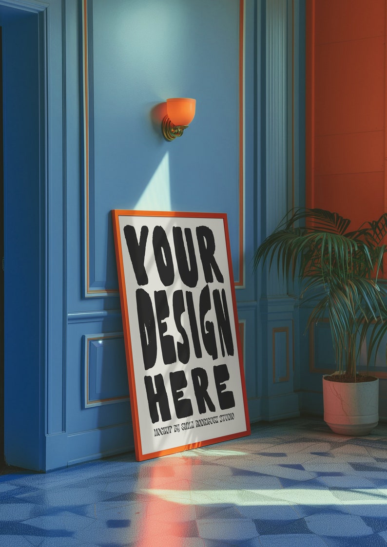 May include: A white poster with a black and orange frame, leaning against a blue wall. The poster says "YOUR DESIGN HERE" in large, black, stylized letters. The poster is in a room with a light blue wall, a light orange wall, and a potted plant.