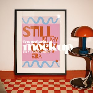 Puede incluir: Impresión enmarcada con el texto "STILL IN MY ERA" en naranja y blanco sobre un fondo rosa. La impresión tiene líneas onduladas azules y está en un marco negro. Una alfombra a cuadros rojos y blancos está en primer plano.
