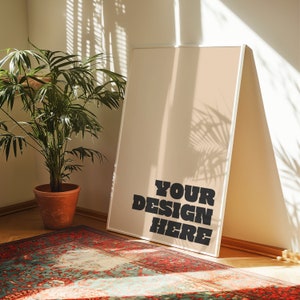 以下が含まれることがあります： 白いフレームにベージュの背景、黒い文字で「YOUR DESIGN HERE」と書かれている。フレームは白い壁に立てかけられており、隅には緑の植物がテラコッタの鉢植えに入っている。床には模様のある敷物が敷かれている。