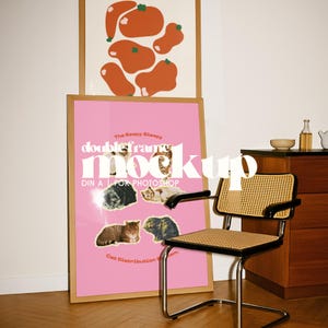 Op de afbeelding: Een roze poster met kattenafbeeldingen en de tekst "The Eeepy Bleepy doubleframe mock up", in een houten lijst. Daarboven een ingelijste abstracte kunstprint met oranje vormen. Een mid-century moderne stoel met een geweven zitting en rugleuning staat op de voorgrond.