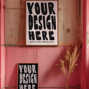May include: Two framed posters with the text "YOUR DESIGN HERE" in black lettering on a white background. The posters are hanging on a pink wall with a dried grass plant in a vase in the foreground.