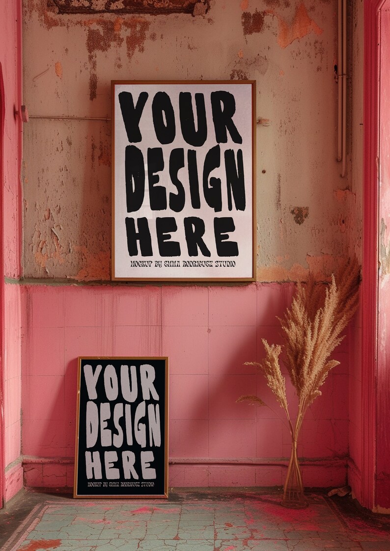 May include: Two framed posters with the text "YOUR DESIGN HERE" in black lettering on a white background. The posters are hanging on a pink wall with a dried grass plant in a vase in the foreground.