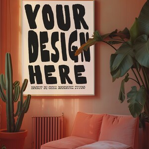 May include: A pink living room with a large framed poster that says "YOUR DESIGN HERE" in black lettering. A cactus plant is in a terracotta pot on the left side of the image. A pink sofa is on the right side of the image.
