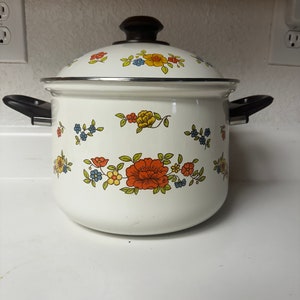 May include: White enamel pot with a floral design in shades of orange, yellow, blue, and green. The pot has a black lid with a wooden knob and two black handles.