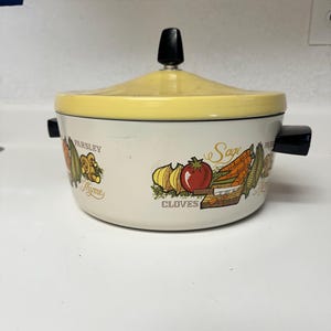 May include: A round, off-white enamelled metal casserole dish with a yellow lid and black handles. The exterior features colourful illustrations of vegetables and herbs, with the printed text labels Parsley, Thyme, Sage, and Cloves visible.