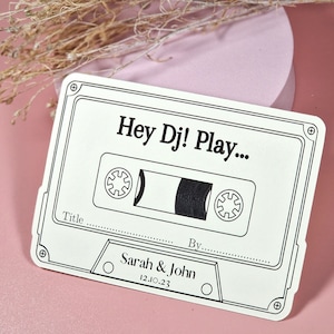 May include: A white cassette tape card with black text that reads "Hey Dj! Play..." with lines for "Title" and "By" and the text "Sarah & John 12.10.23" below the cassette tape graphic.