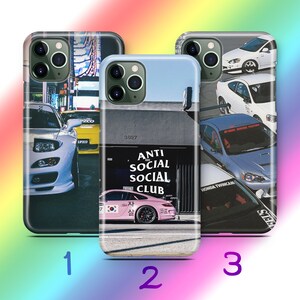 Puede incluir: Tres fundas para teléfono con diseños de coches. Una funda muestra un coche blanco en un entorno urbano, otra un coche rosa con el texto "ANTI SOCIAL SOCIAL CLUB", y la tercera varios coches.