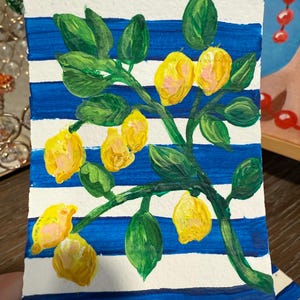 May include: A small, rectangular painting featuring a branch of lemons. The lemons are yellow with pink highlights, set against a blue and white striped background. Green leaves and stems complete the artwork.