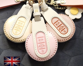 May include: Three leather keychains with a white background and pink, peach, and cream colored accents. Each keychain has a different design of a car key with a small car icon and the letter "B" on it. The keychains are attached to gold rings and a gold chain.