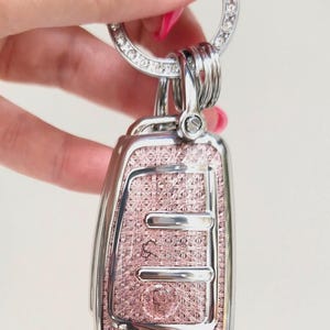 May include: A decorative car key fob with a pink patterned face and a silver-colored frame. The key fob is attached to a silver key ring adorned with clear rhinestones. The key fob has a button and several silver-colored key rings.
