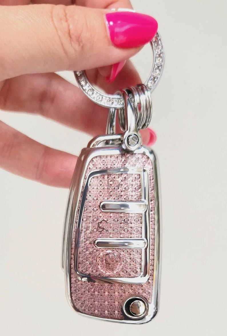 May include: A decorative car key fob with a pink patterned face and a silver-colored frame. The key fob is attached to a silver key ring adorned with clear rhinestones. The key fob has a button and several silver-colored key rings.