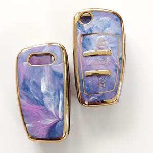 May include: Two car key fobs with a marbled design in shades of purple, blue, and white. The key fobs have a gold-colored trim. One fob has a key ring hole, and the other has buttons for car functions.